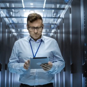 IT Technician Walks Through Rows of Server Racks in Data Center. Simultaneously He Works on a Tablet Computer.